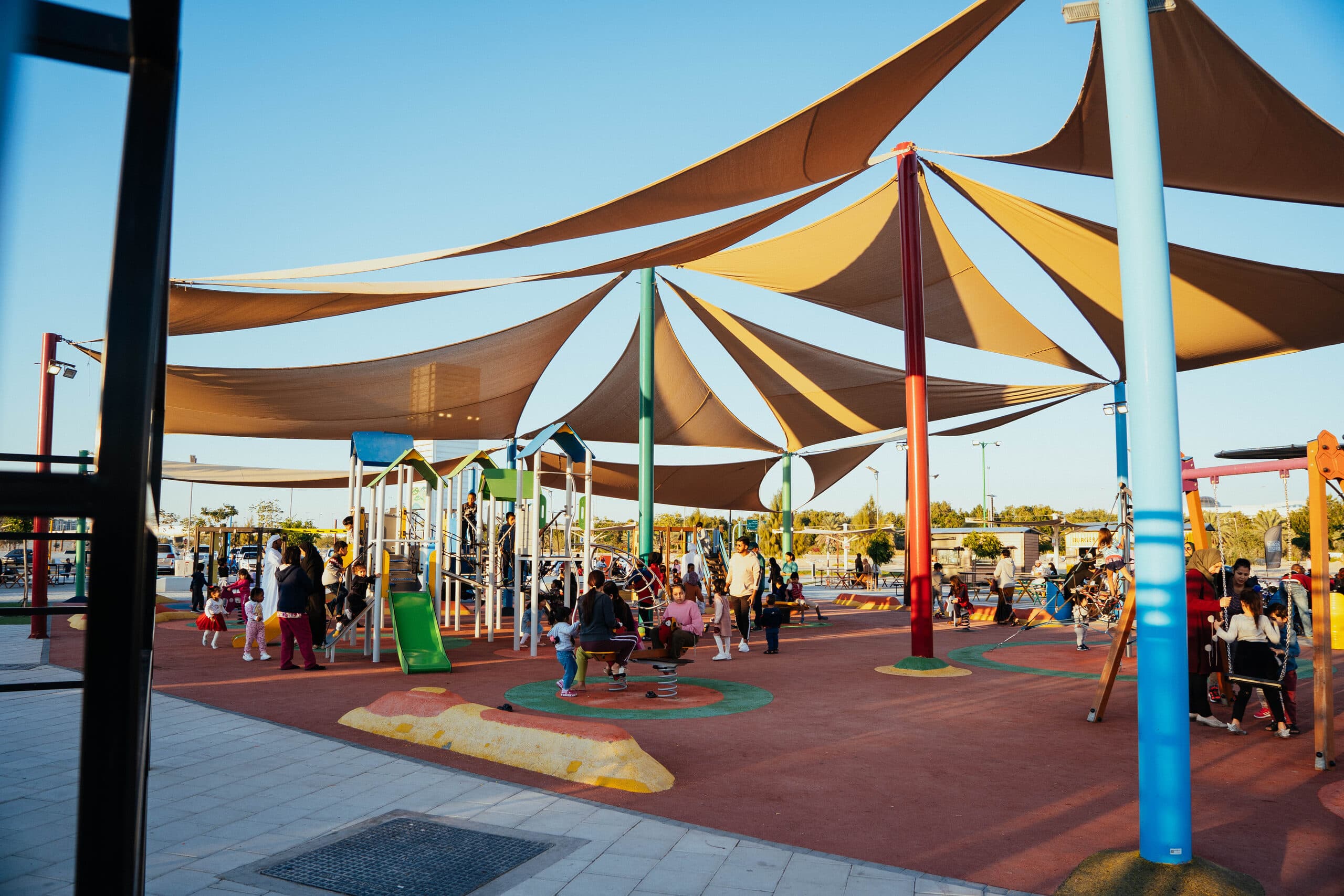 Shade sails over a playground in Masdar Park, families on the benches