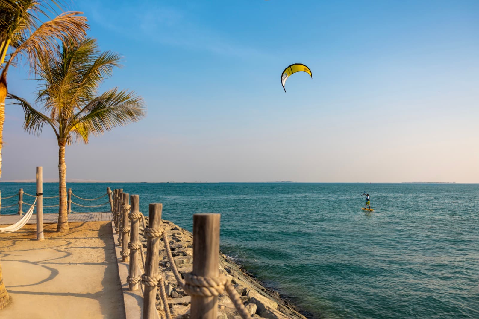 Palms and a rope-railed boardwalk at Yas Beach, a kite surfer offshore